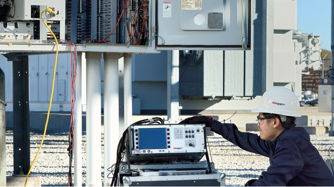 Dynamo field services technician using specialized tools to test electrical system. The location is a substation. Employees are wearing a hardhats gloves and eye protection.