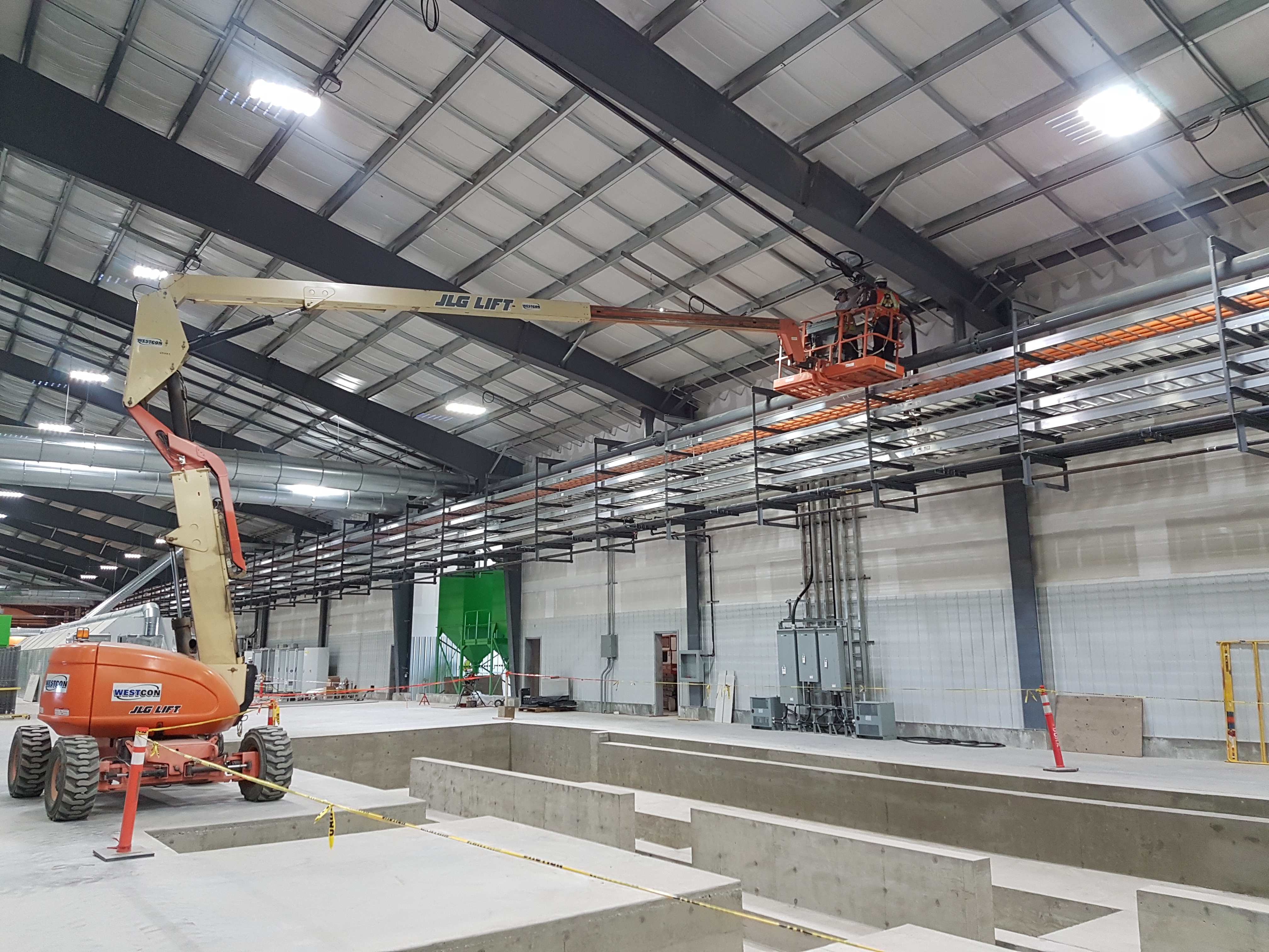 Dynamo employees on a boom lift high in the rafters runing electrical wire for an industrial building. All two employees are wearing Personal Protective safety equipment and fall arrest harasses