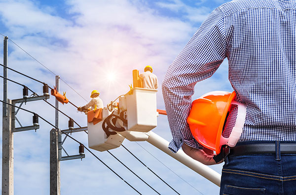  Two employees are in the bucket of a boom truck using High Voltage Hot Stick on the power line as a supervisor watches. The employees are wearing a hard hats, gloves and protective eyewear.