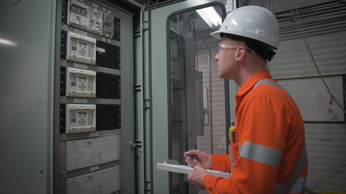 Dynamo electrician taking notes at an electrical panel in preparation for protective relay testing. The employee is wearing a hard hat coveralls and protective eyewear.