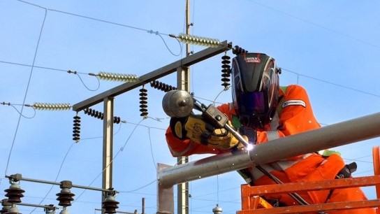 Dynamo welder in the field performing aluminum bus welding. The employee is wearing a welding helmet, gloves, coveralls and fall arrest harness.
