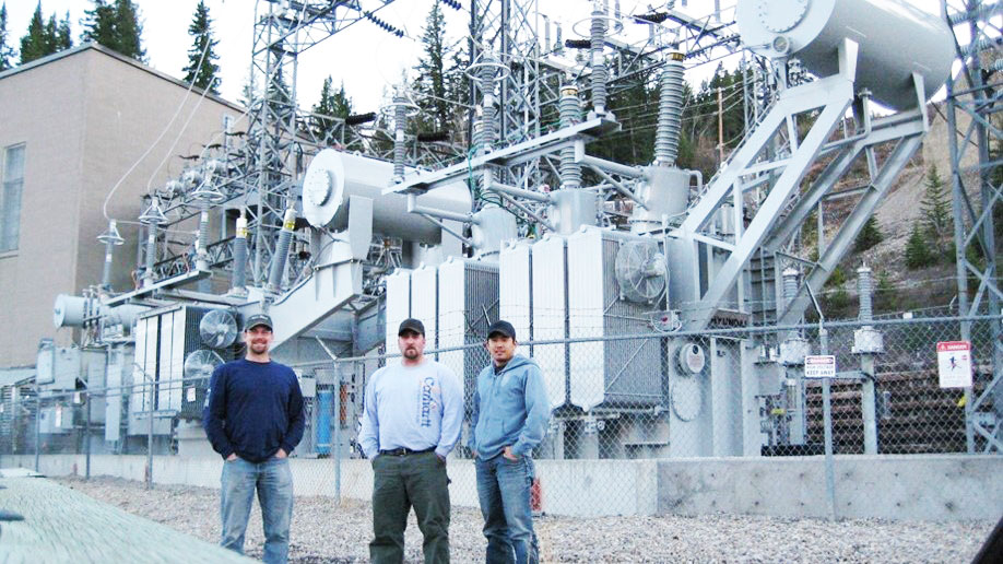 Three Dynamo employees standing in front of an electrical substation. The location is in a rocky forest. The employees are proud of the service they provided.ee Dynamo employees standing in front of an electrical substation. The location is in a rocky forest. The employees are proud of the job they completed.