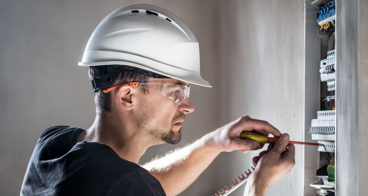  Dynamo electrician installing breakers into a panel with a screwdriver. The employee is in wearing a hard hat and protective eyewear.