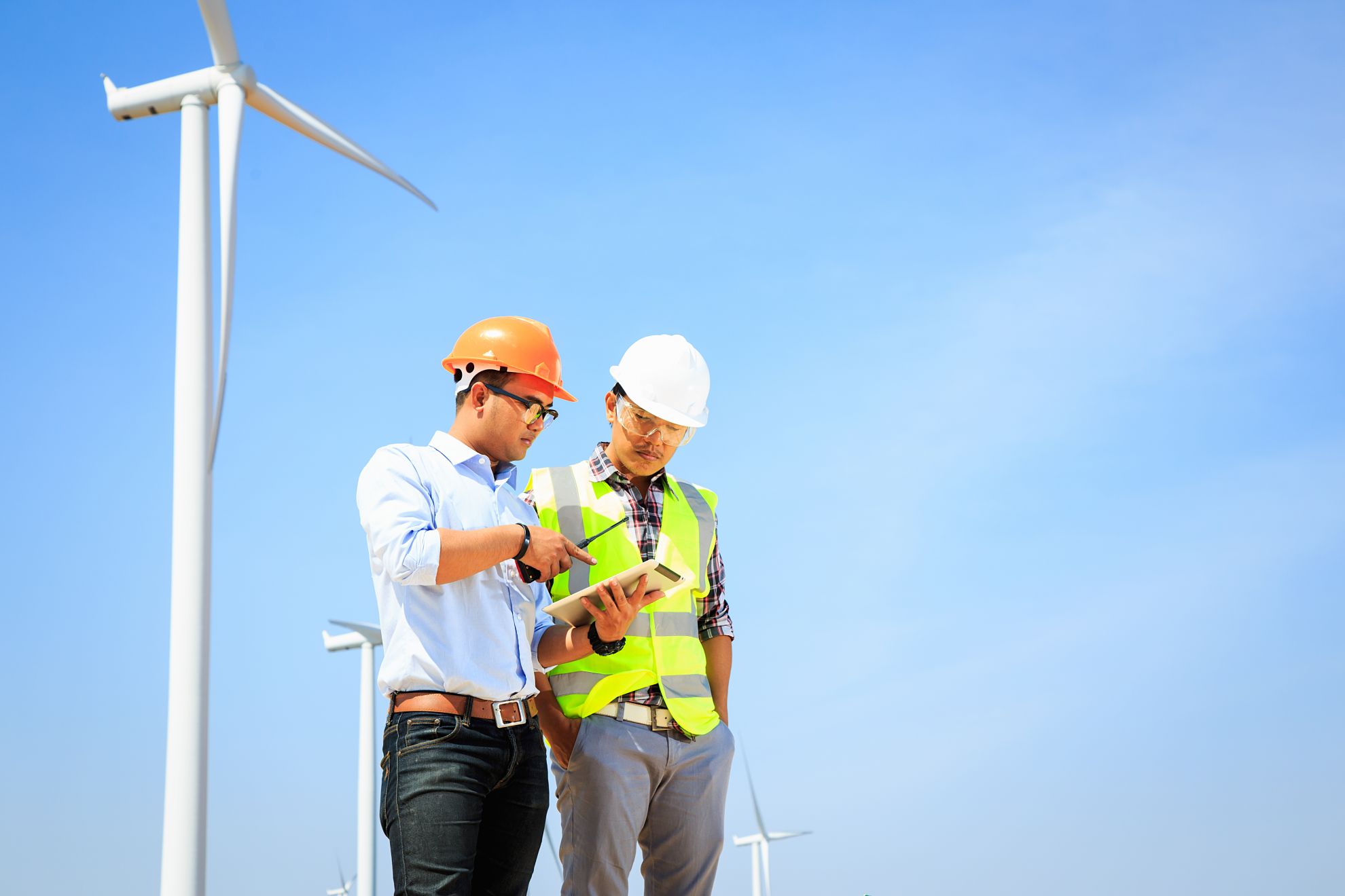  Two Dynamo engineers review information on a tablet. They are in a wind turbine field and are both wearing personal protective equipment.