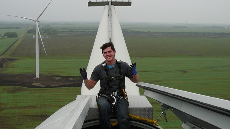 Dynamo employee sitting on the top of a wind turbine. The location is a field of wind turbines. The employee is wearing gloves and fall protection harness.